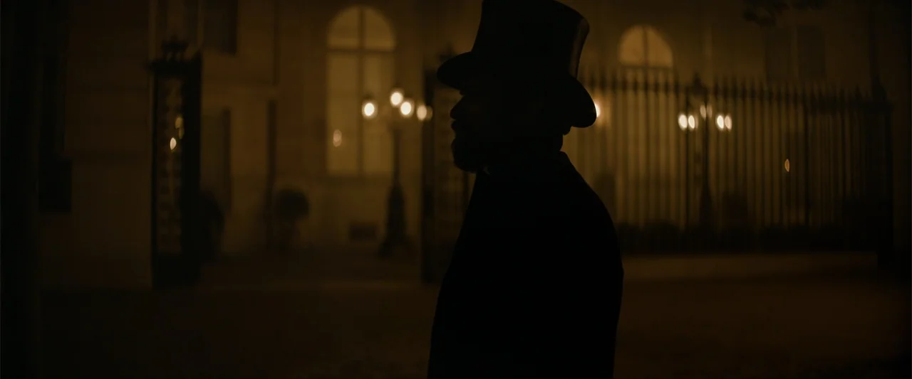 Eiffel (2021) movie still: medium — A dark city street features an ornate metal gate illuminated by distant lamp lights. A man in a top hat stands…