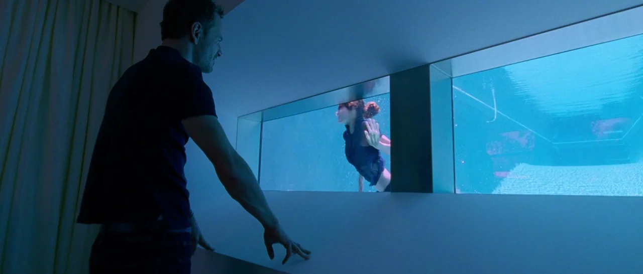 Song to Song (2017) movie still: medium — A modern room looks into a blue-lit pool through horizontal glass viewports. A man stands in the room…