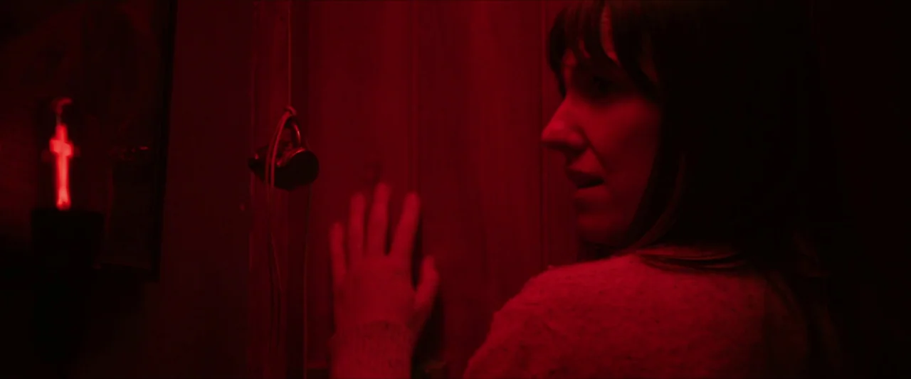 Fréwaka (2024) movie still: medium — A dark, dimly lit corridor features wooden paneling and a glowing red wall ornament. A woman in a knitted…