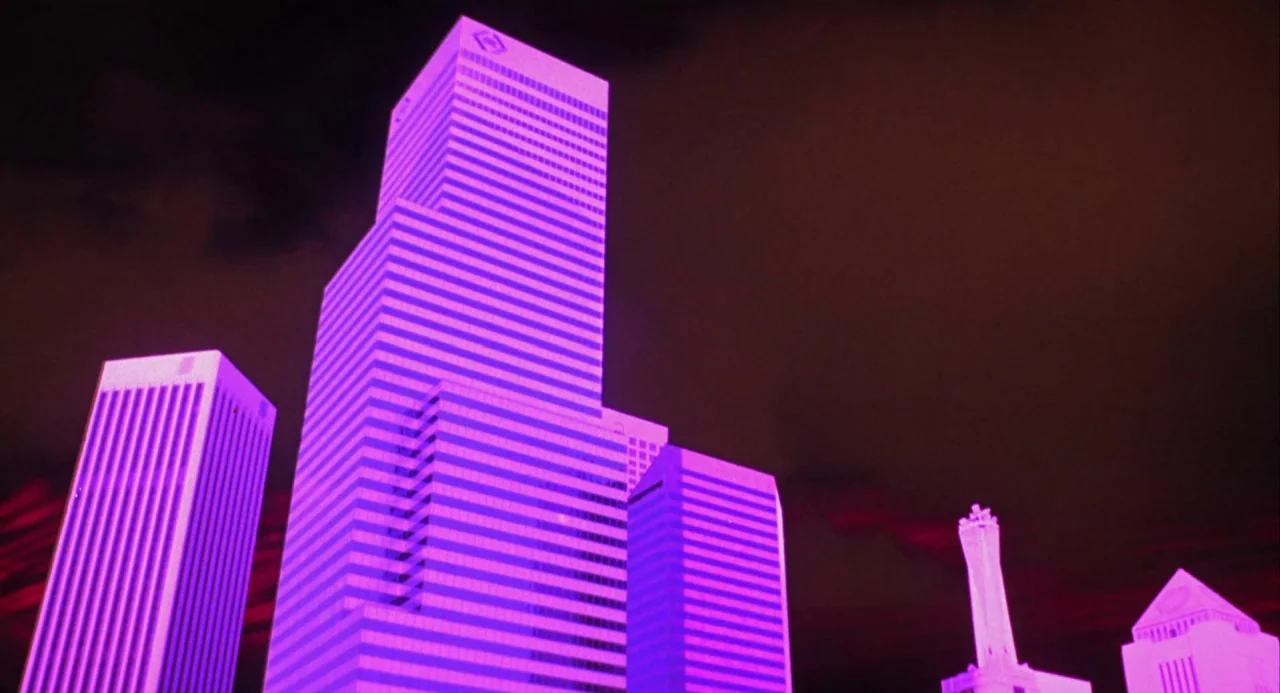 Night of the Comet movie still: establishing — Modern skyscrapers dominate the frame from a low angle in a city center. Intense purple light floods…