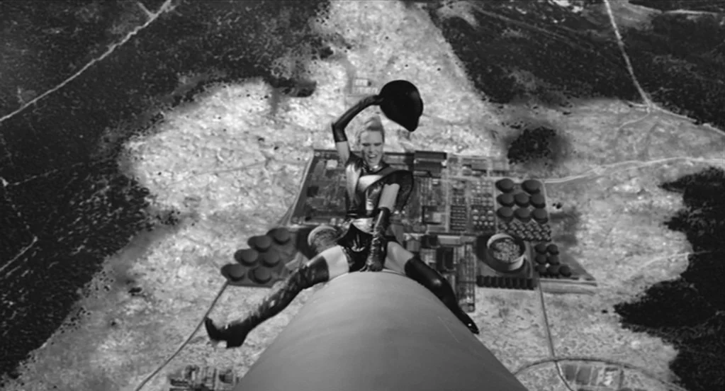 The Congress (2013) movie still: fullbody — A woman sits atop a narrow chimney overlooking an expansive black-and-white industrial landscape. The…