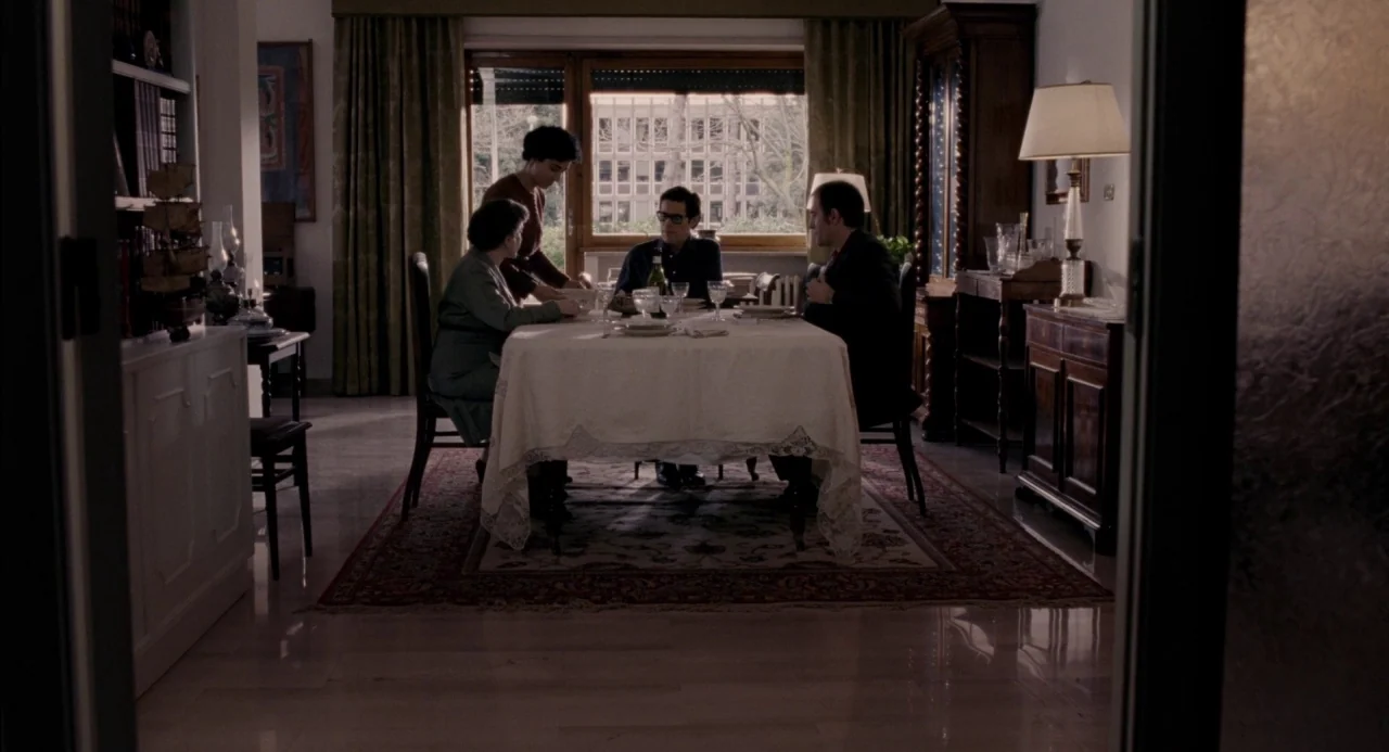 Pasolini (2014) movie still: wide — A well-appointed dining room features a long table set for a family dinner. Four adults sit around the table…