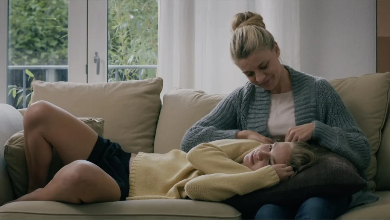Copenhagen (2002) movie still: medium — A sunlit living room with a neutral-toned sofa near a white framed window. An adult woman strokes the hair…