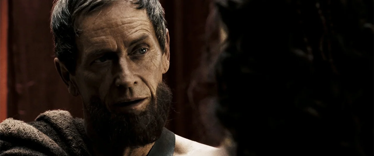 300 (2007) movie still: closeup — A dark indoor space with minimal architectural detail visible in the background. A man with a textured beard and…