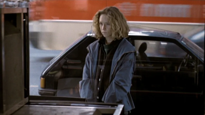 Benny’s Video (1992) movie still: medium — A woman stands behind a glass surface in an urban environment during the daytime. Behind her, a dark car…