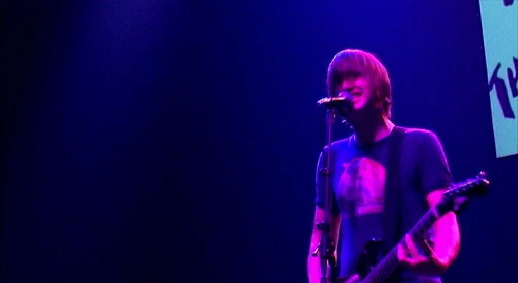 9 Songs (2004) movie still: medium — A musician performs on a dimly lit stage against a dark background. He holds a microphone and plays a guitar…