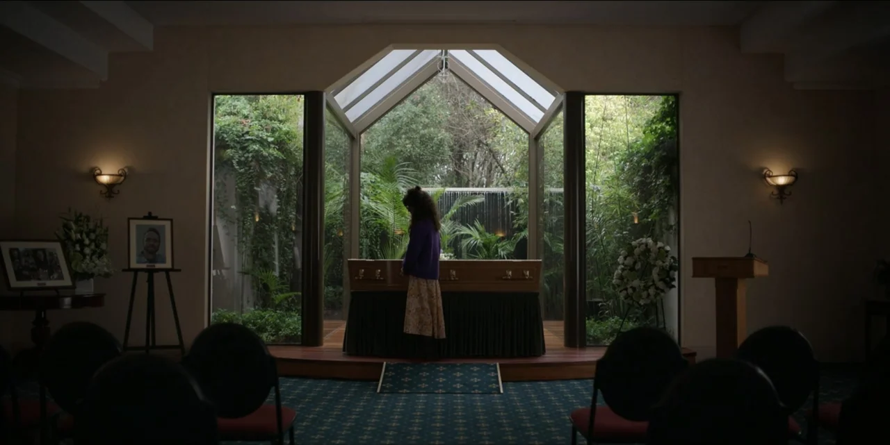 Bring Her Back (2025) movie still: fullbody — A funeral home interior features a central casket positioned before a large glass wall. A woman in a…