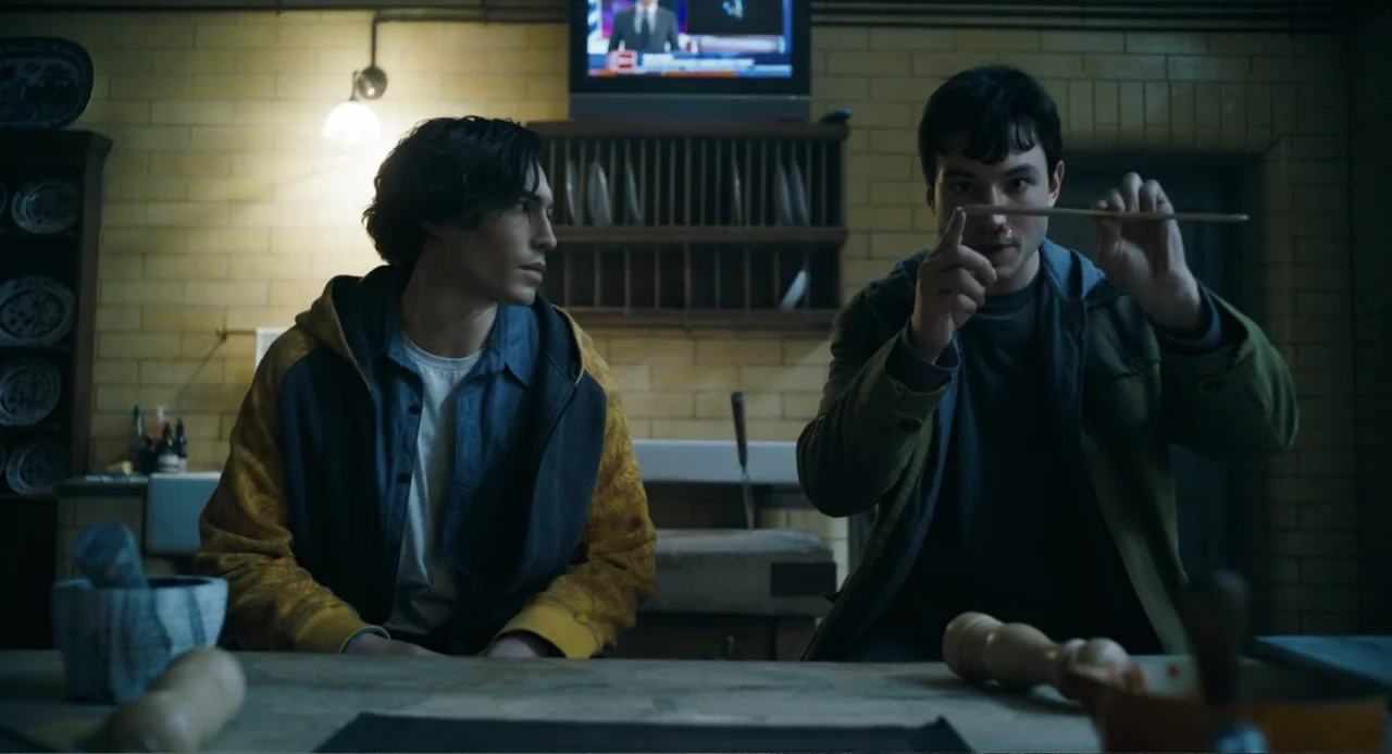 The Flash (2023) movie still: medium — A dimly lit room with yellow-tiled walls features a wooden table and shelving. Two young men sit at the table…