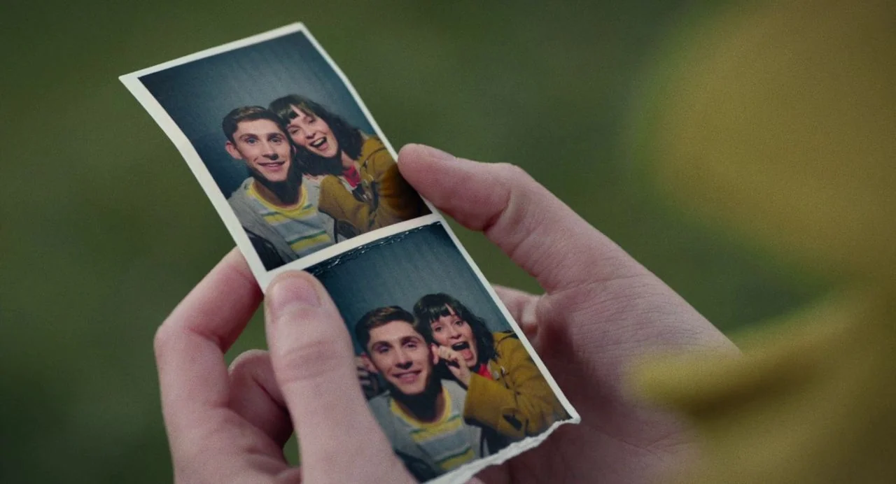 Dating Amber movie still: closeup — A pair of hands holds a double-frame photo booth strip against a green background. The photograph shows a young…