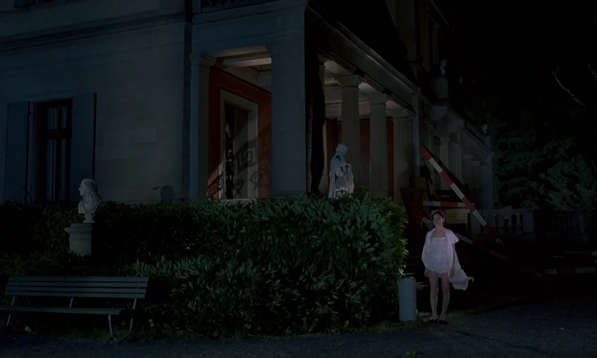 Phenomena (1985) movie still: wide — A classical style mansion features white pillars and statues set against a dark night. A woman wearing white…