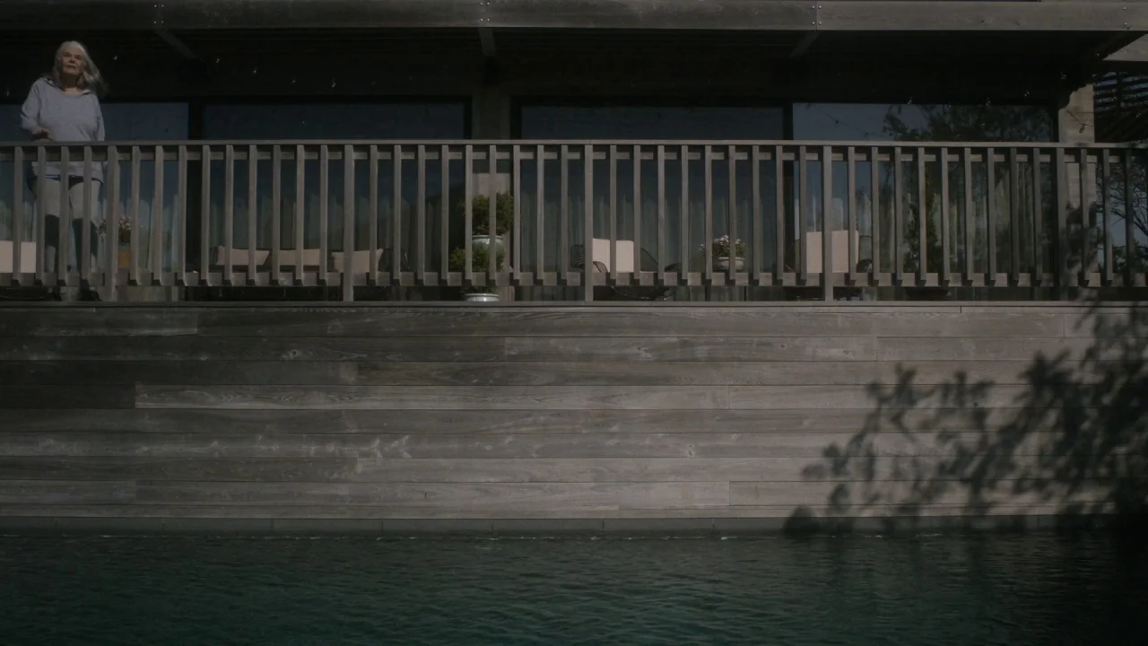 Marjorie Prime (2017) movie still: wide — A modern house deck built from grey wood overlooks a dark pool of water. An elderly woman stands alone on…