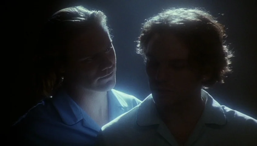 Edward II (1991) movie still: medium — A dimly lit room provides a dark, obscured backdrop for two men. The subjects stand close together while…