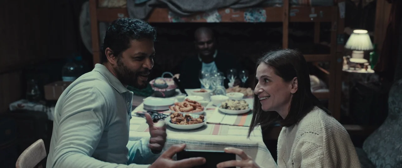 R.M.N. (2022) movie still: medium — A cluttered dining space with wooden furniture and a table covered with various dishes. A man and a woman face…