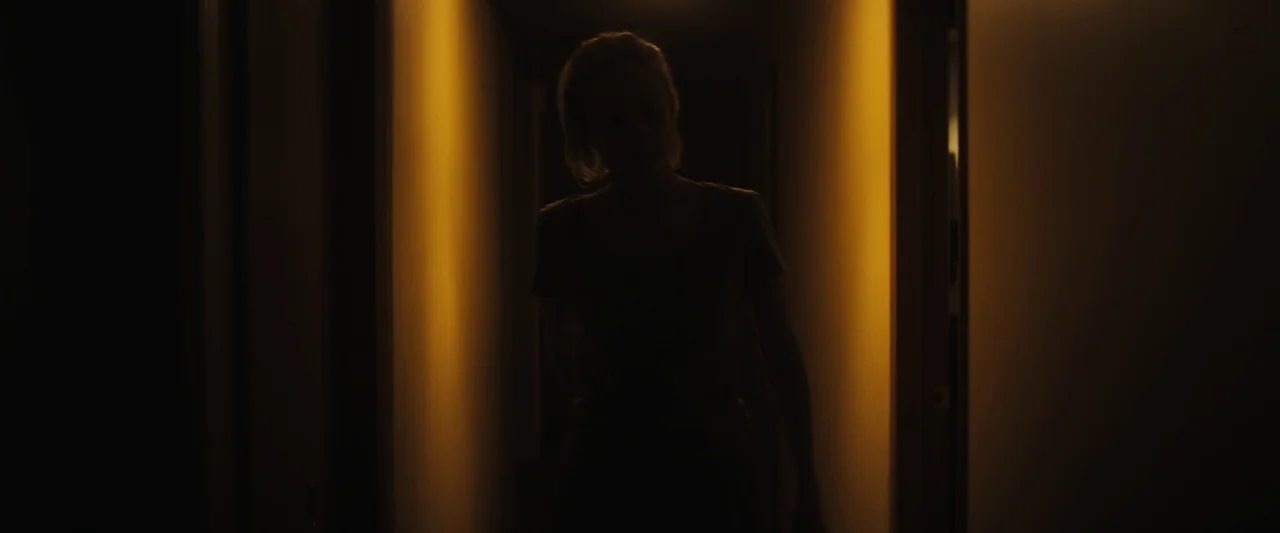 The Grudge (2020) movie still: medium — A dark hallway is illuminated by a thin strip of warm light. A person stands in the center of the frame as a…