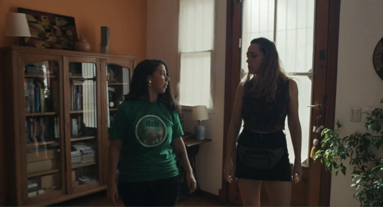 Belén (2025) movie still: medium — A residential room features a tall wooden glass-fronted cabinet filled with books and items. Two young women…