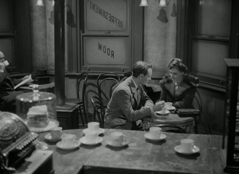 Brief Encounter (1945) movie still: medium — A 1940s refreshment room features wooden furniture, curved chairs, and a glass cake cover. A man and a…