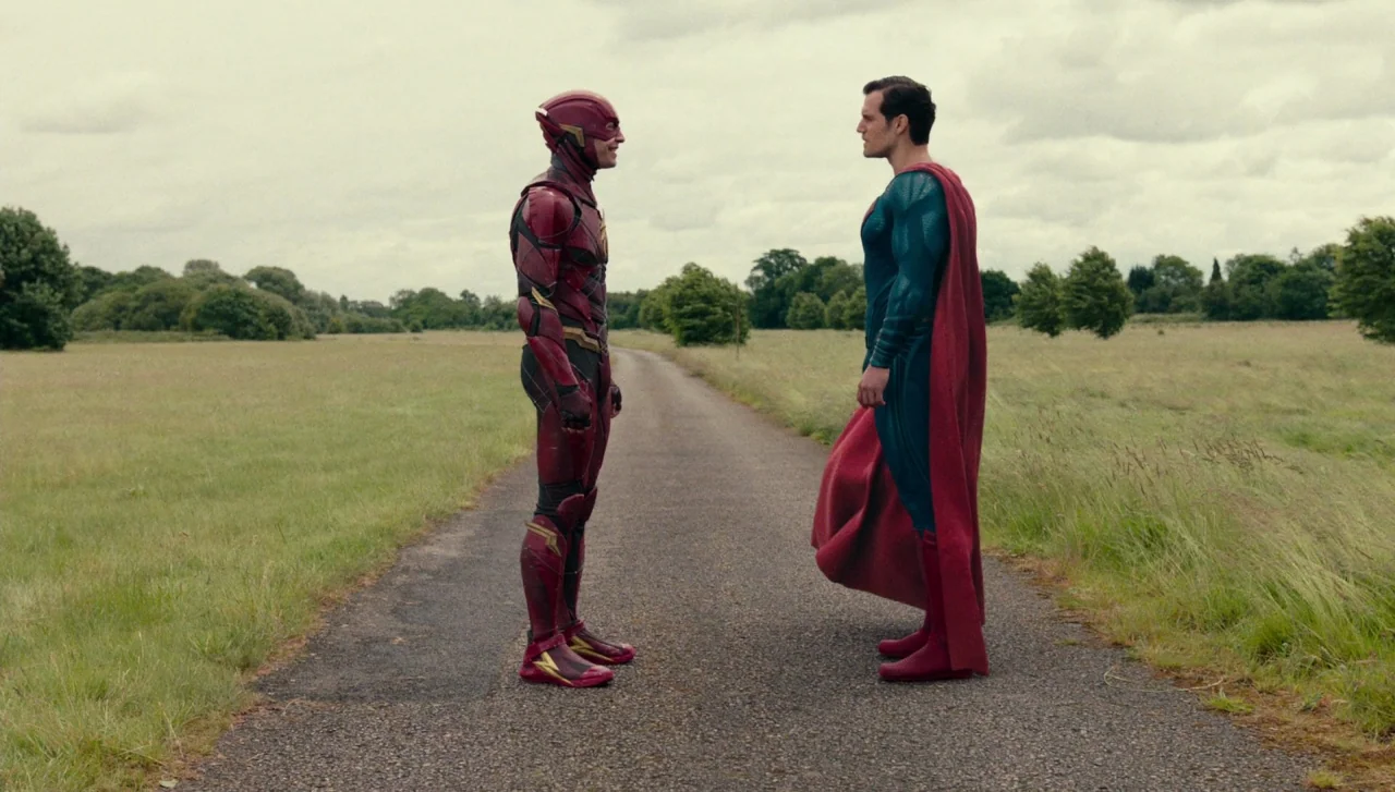 Justice League (2017) movie still: fullbody — A rural paved road cuts through a wide expanse of overgrown green fields. The Flash and Superman stand…