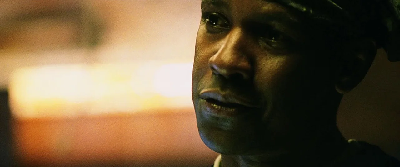 Man on Fire (2004) movie still: closeup — A close-up shot focuses on a man in a dimly lit indoor space. He gazes off-camera while soft ambient light…