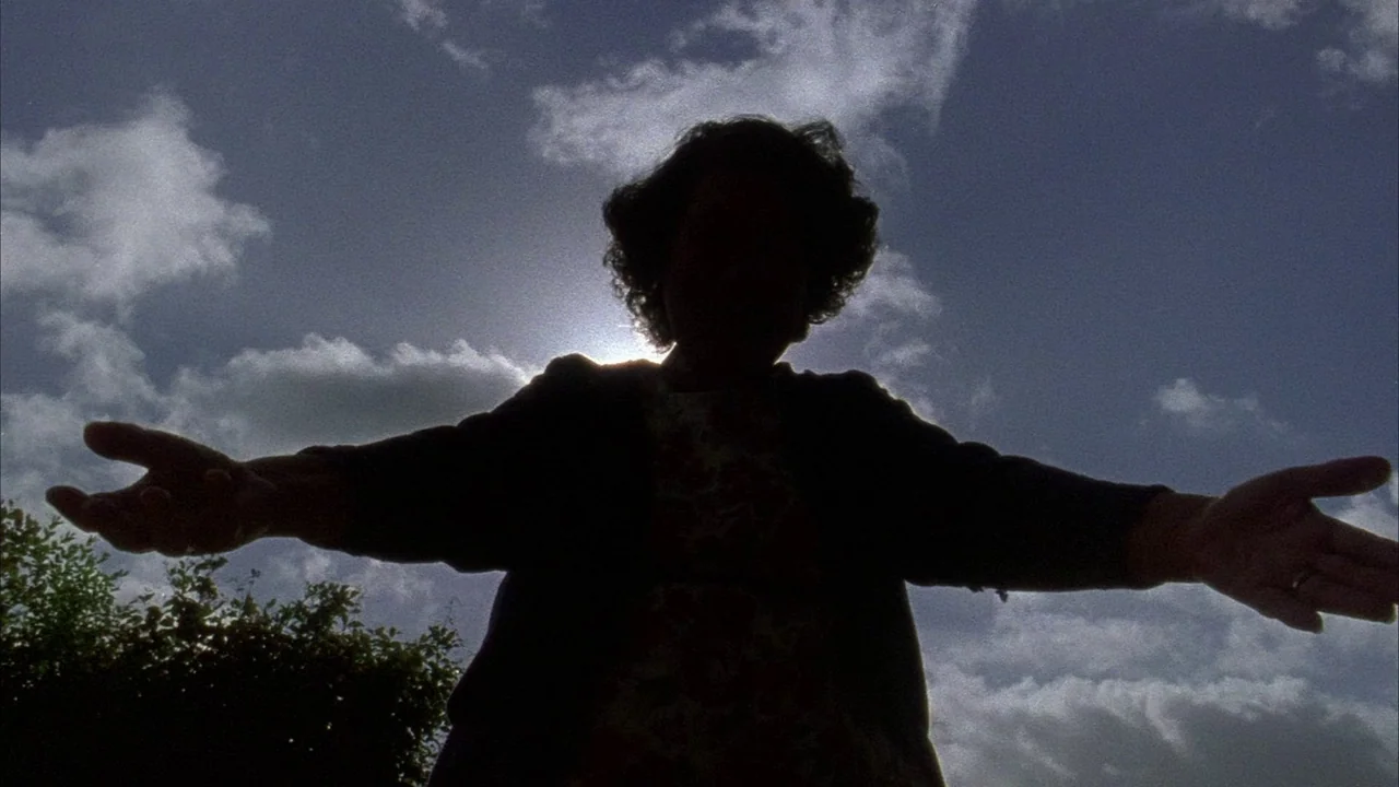 An Angel At My Table (1990) movie still: medium — A person stands against a bright, cloudy sky with sun rays partially obscured. The subject holds…