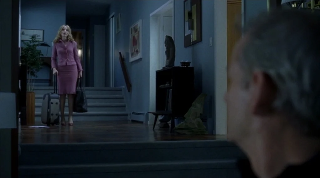 Broken Flowers movie still: wide — A residential hallway with blue walls and a staircase leading to a landing. A woman carrying luggage stops at the…