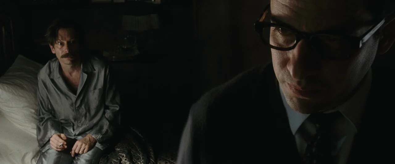 Chicken With Plums (2011) movie still: medium — A dark hotel room interior at night with a man in the foreground in focus. A man in pajamas sits on…