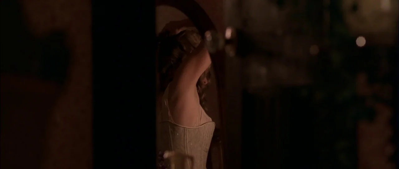 Open Range (2003) movie still: medium — A dimly lit room contains a large, dark-framed standing mirror. A young woman in a corset turns to fix her…