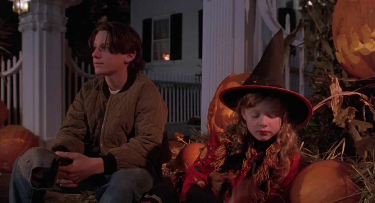 Hocus Pocus (1993) movie still: medium — A residential house porch decorated with straw bales and large orange pumpkins at night. A teenage boy sits…