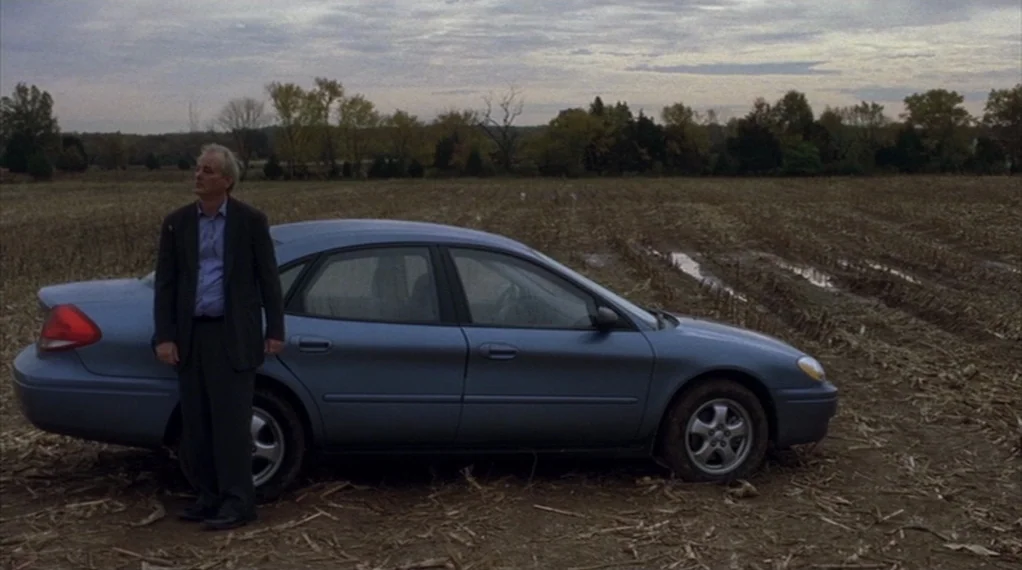 Broken Flowers movie still: wide — A man stands next to a blue sedan in a wide, harvested brown field. The sky is overcast and the ground is muddy…