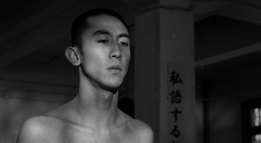Mishima: A Life in Four Chapters (1985) movie still: medium — A dark indoor space with a concrete pillar featuring Japanese calligraphy on a sign. A…
