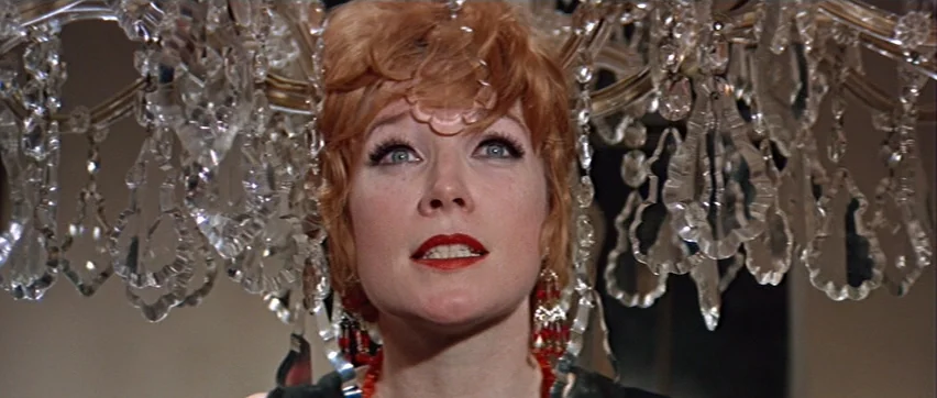 Sweet Charity (1969) movie still: closeup — A woman stands directly beneath a large, ornate crystal chandelier indoors. The woman looks upward with…