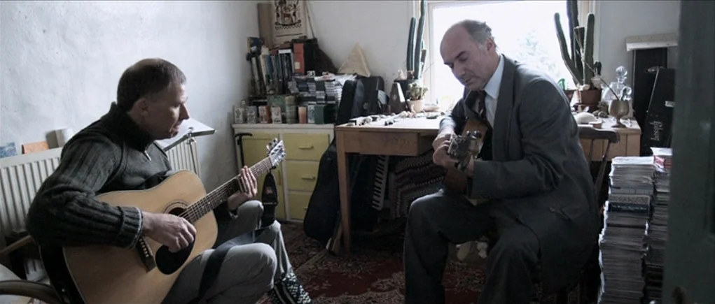 Down Terrace (2009) movie still: medium — A cluttered room features a wooden desk, stacks of books, and cactus plants. Two men sit on chairs facing…