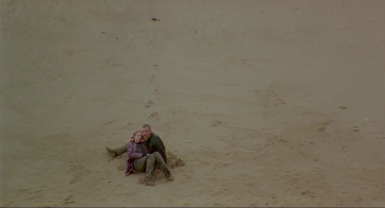 Romper Stomper (1992) movie still: wide — An expansive, empty sand dune stretches across the frame under an overcast sky. A man and woman sit…