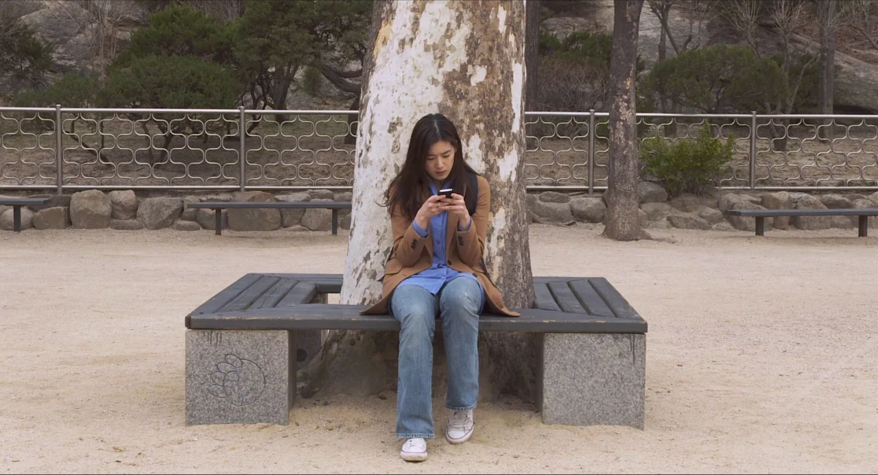 Nobody’s Daughter Haewon (2013) movie still: medium — A woman sits on a square bench encircling a large tree in a park. She is looking down at a…