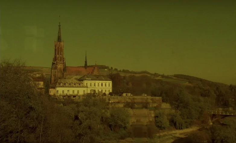 The Double Life of Véronique (1991) movie still: establishing — A tall gothic-style church stands next to a large building atop a grassy hill. The…