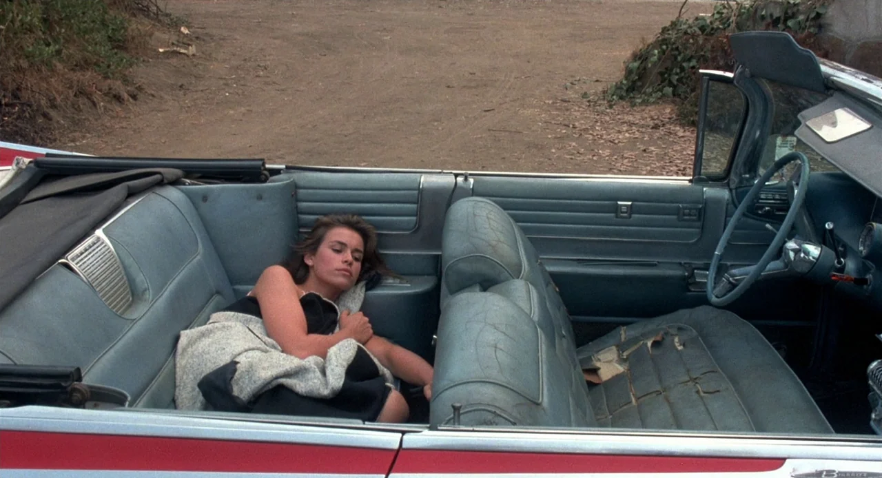 Breathless (1983) movie still: medium — A vintage American convertible with cracked light blue leather seats parked on dirt ground. A young woman…