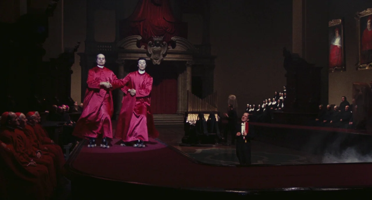 Roma (1972) movie still: wide — A large, dim religious hall features red-carpeted platforms and framed paintings on walls. Two men dressed in red…