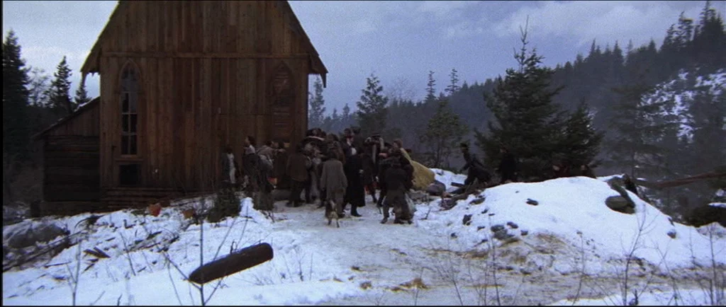 McCabe & Mrs.Miller (1971) movie still: wide — A rustic wooden building stands on a snow-covered hill surrounded by coniferous trees. A small group…