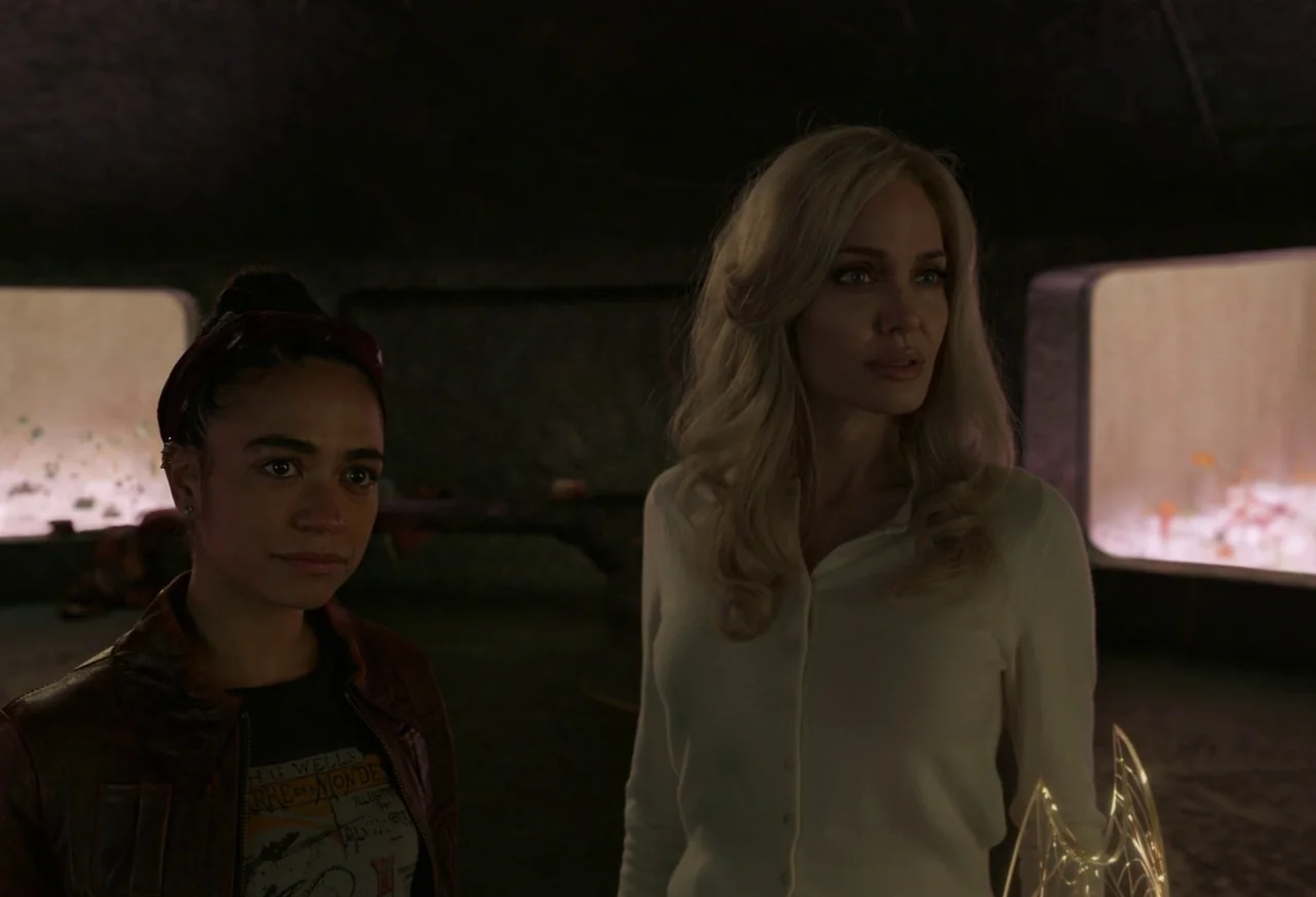 Eternals (2021) movie still: medium — A dark exhibition room features glowing glass cases holding curated artifacts along the back wall. Two women…