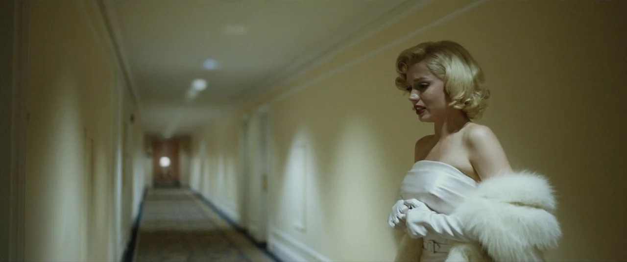 Blonde (2022) movie still: medium — A long hotel corridor features pale yellow walls and neutral carpeting under recessed lighting. A woman in a…
