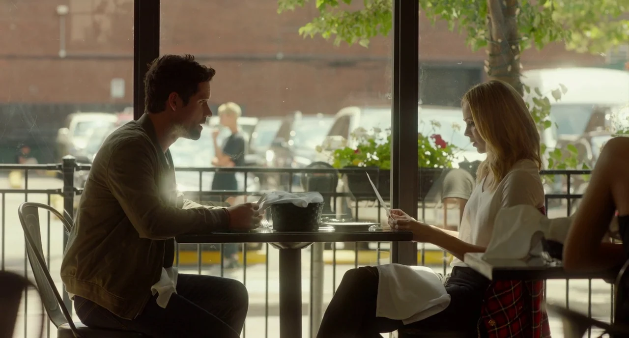 Rabid (2019) movie still: medium — A man and woman sit at a square table inside a brightly lit restaurant. They face each other while reading menus…