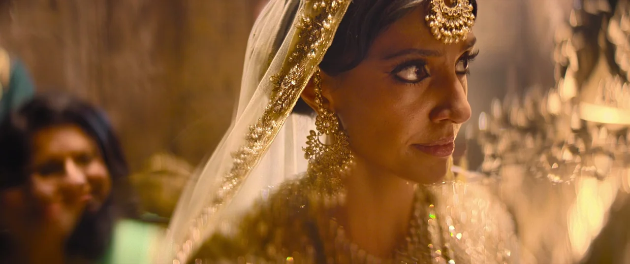Polite Society (2023) movie still: closeup — A woman wears traditional Indian wedding attire with intricate gold jewelry and a veil. She glances to…
