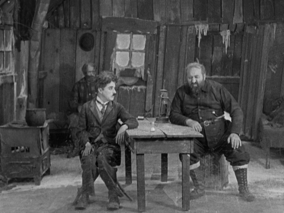 The Gold Rush (1925) movie still: fullbody — A rustic log cabin interior features rough wooden walls and a simple plank table. Two men sit at the…