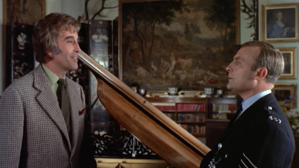 The Wicker Man movie still: medium — A traditional room features a grand piano in the foreground and a large tapestry. A man in a tweed blazer…