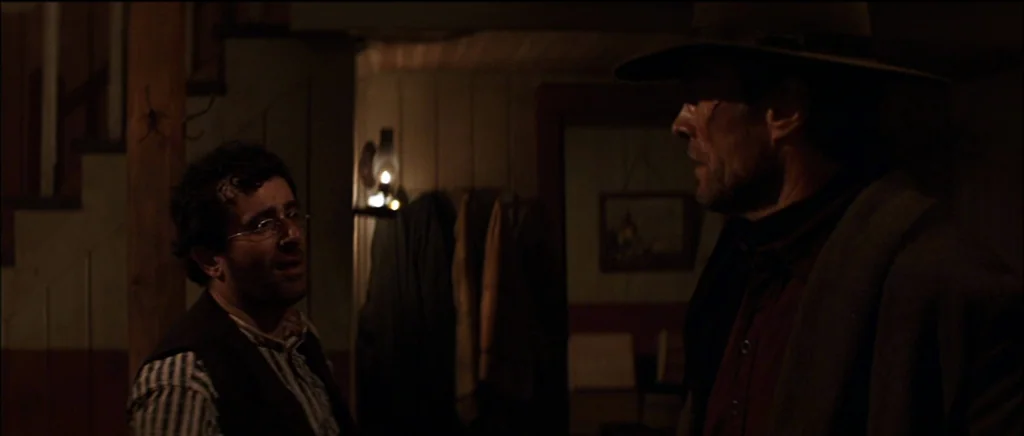 Unforgiven (1992) movie still: medium — A dark wooden room lit by a faint lamp in the background. A man in glasses addresses a man wearing a cowboy…