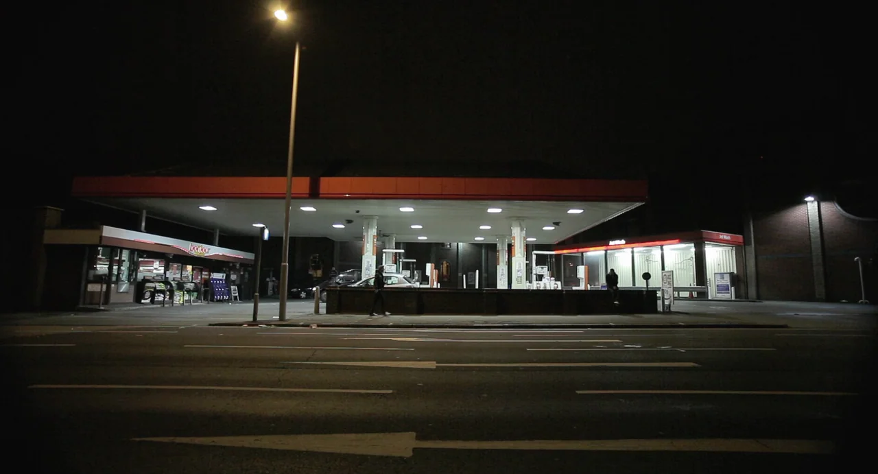 Weekend (2011) movie still: wide — A deserted gas station stands beside a multi-lane road at night. Bright overhead canopy lights illuminate the…