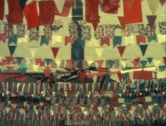 Glitterbug (1994) movie still: establishing — Multiple rows of small colorful bunting and Union Jack flags hang over a street. The fabric displays…