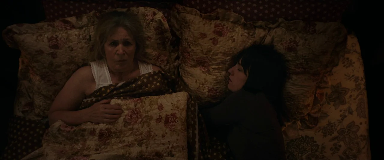 Fréwaka (2024) movie still: medium — An overhead shot of two women resting in a bed with floral-patterned linens. An older woman is awake while a…