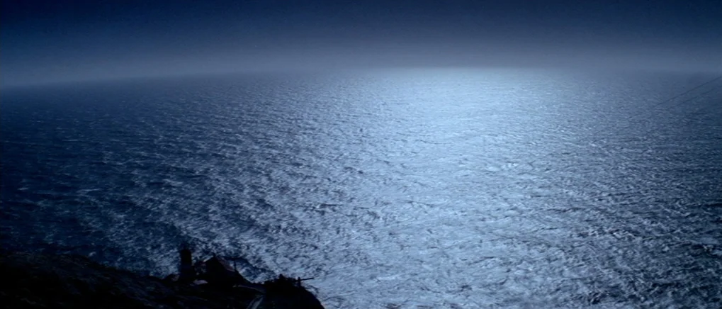 The Fog (1980) movie still: establishing — A dark rocky cliff with a small structure sits beside a vast ocean. Moonlit water ripples under a heavy,…
