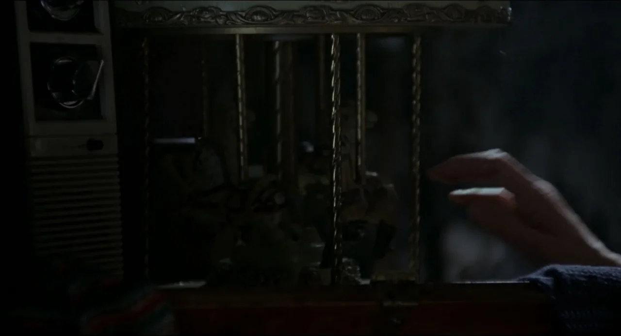 The Ring Two (2005) movie still: closeup — A dimly lit room contains a vintage miniature carousel set beside an old television. A human hand extends…