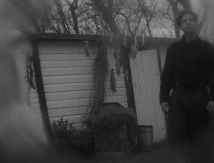 Tales from the Gimli Hospital (1988) movie still: medium — A white wooden shed sits under bare trees with fishing nets hanging outside. A young man…